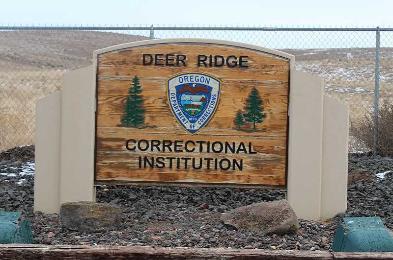 A wooden sign reading Deer Ridge Correctional Institution sits in front of a chainlink fence with barren hills in the background.