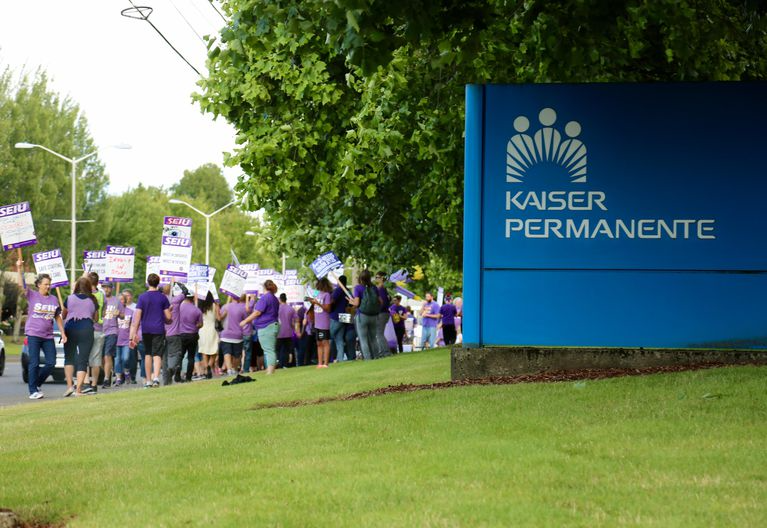 A large group of people wearing purple shirts and holding labor union signs stand in front of Kaiser building.