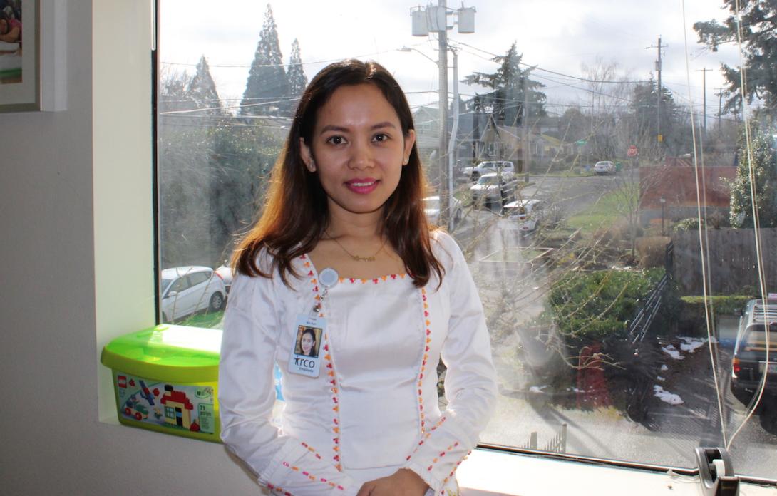 A young woman with tan skin, dark brown hair stands in front of a window wearing a white shirt.