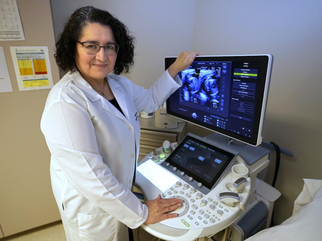 A woman in a white doctor's coat stands in front of a medical computer terminal.
