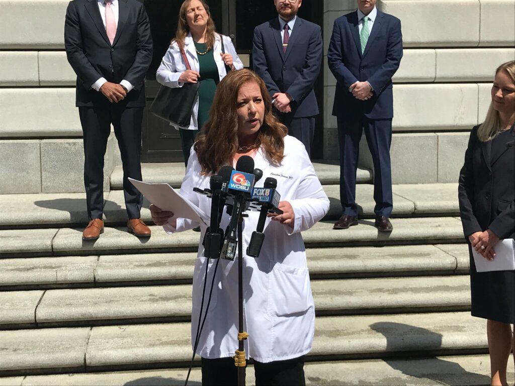 A woman in a doctor's white coat speaks on a courthouse steps.