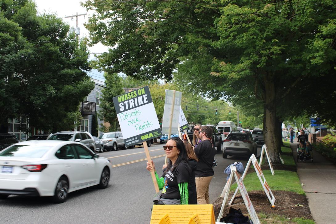 people hold signs along a street as cars pass by, one sign says 'people not profits'