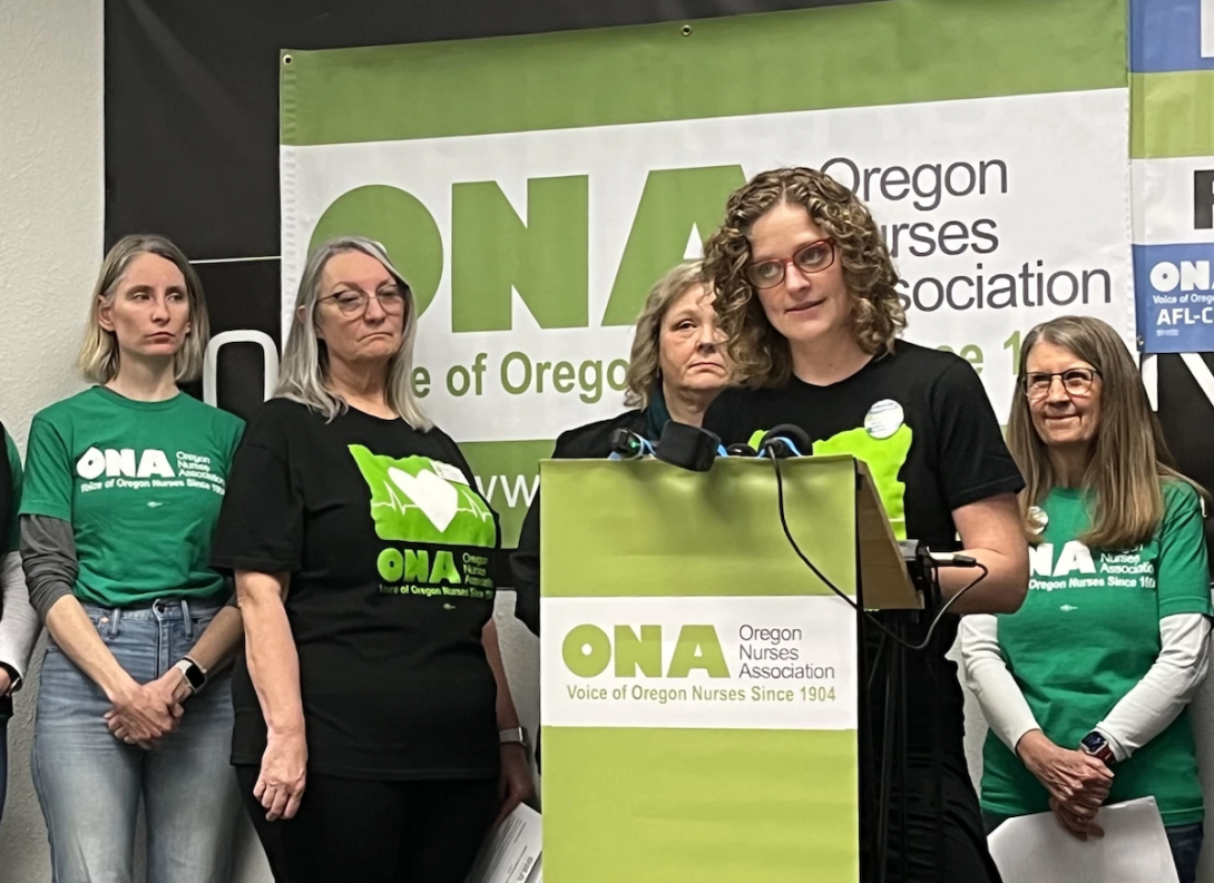 home care nurses presser A group of women stand, as one speaks at a lectern.