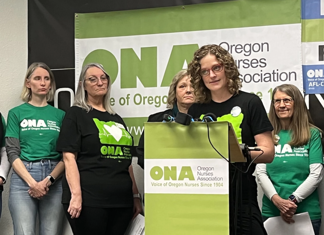 A group of women stand, as one speaks at a lectern.