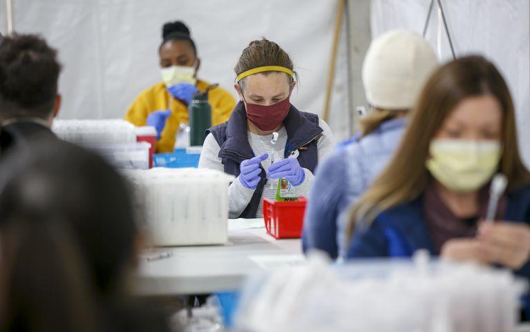 A woman wearing a mask prepares a syringe for vaccination injection at a table under a tent in a parking lot.