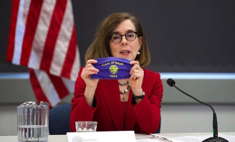 Gov. Kate Brown holds a mask with Oregon's state crest pictured on it as she sits behind a desk.