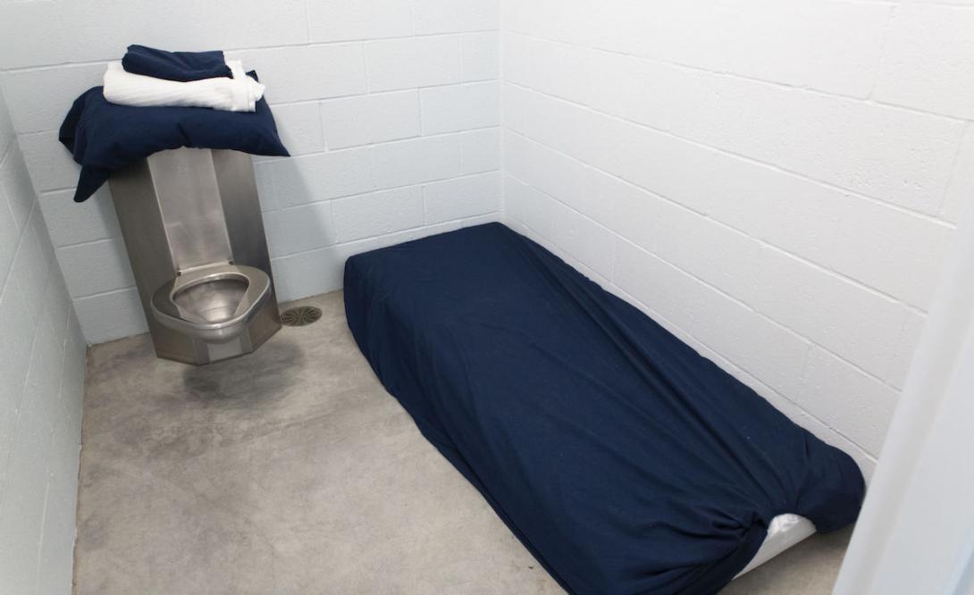A room resembling a jail cell, with cement walls, a cot and metal toilet. 