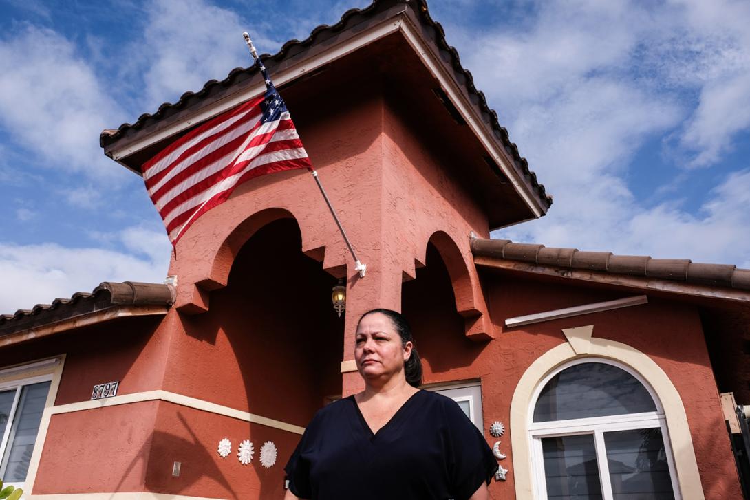 A middle-aged Hispanic woman standing in front of a stucco house displaying an American flag flapping in the wind.