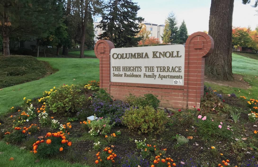 A sign in a bed of flowers reads The Heights at Columbia Knoll, with a building in the background on well kept landscape.