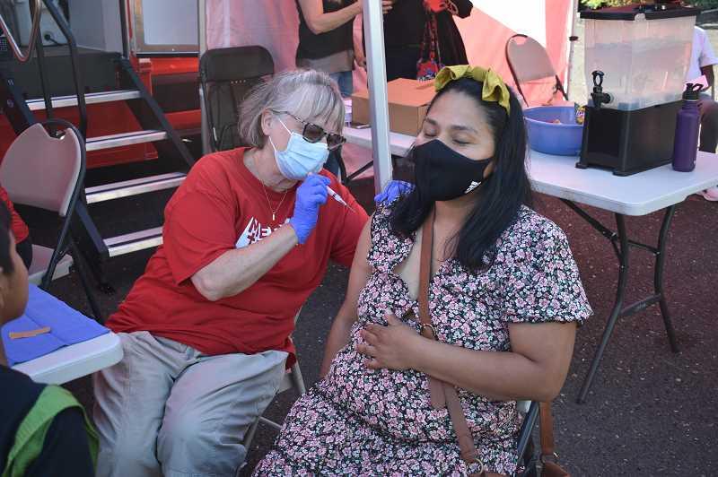 A woman with closed eyes sits in a chair while another woman holds a needle inches away from her right arm.