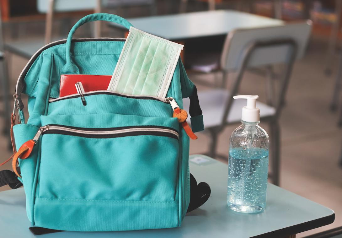 Backpack on a desk with a medical mask sticking out of its pocket and hand sanitzer